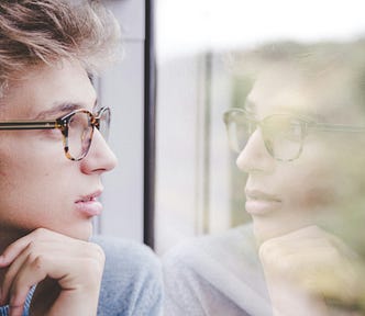 young man with glasses looking out the window, in which we see his reflection.