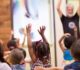 This image features a group of young children in a classroom, with several of them raising their hands high. In the background, an adult with gray hair is also raising his hands, suggesting he might be leading the group in an activity. The focus is on the children, who are engaged and appear to be having fun.