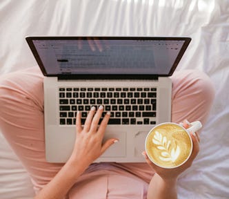 Overhead view of a woman sitting cross-legged in pink leggings, holding an artistically on-point cup of coffee and typing on a laptop. She also seems to be sat on a white sheet so maybe she’s on her bed, or maybe she’s having a picnic, who knows?