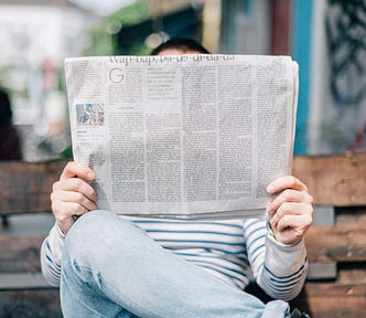 A person sites on a park bench reading a newspaper, holding the paper so that it blocks their entire face.