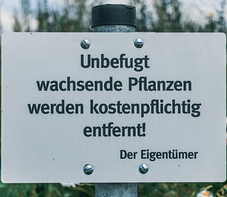 White sign on metal pole in flowery summer garden, in German, reading “Unbefugt wachsende Pflanzen werden kostenpflichtig entfernt! Der Eigentümer”