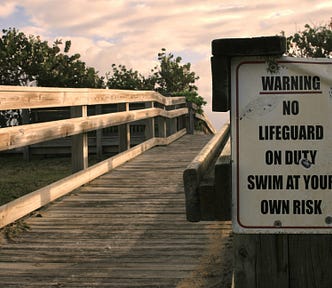 A small sign set up at the beginning of a boardwalk leading to a beach. It reads: Warning: No lifeguard on duty, swim at your own risk.