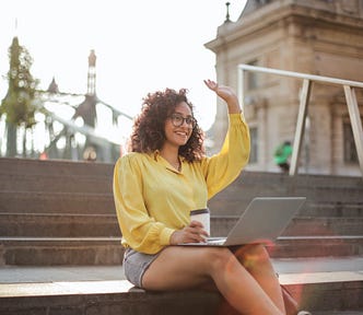A woman of color is sitting on outdoor steps, smiling and waving. Her laptop and a cup of coffee are in her lap.