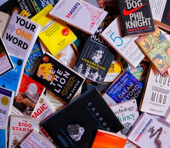 A pile of self-help books strewn across the floor. Some of the visible titles include “Your One Word,” “Shoe Dog,” and “The Code To An Extraordinary Mind.”