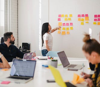 A group of people in a workshop session. A woman with long hair is standing at the front of the room pointing at a wall of post it notes. The other people are sitting around the table with laptops.