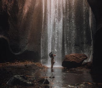 A lowly dim long shot of a woman with a hat in front of a thin sheet of waterfall in front and surrounded by tall rock and low light as a metaphor of insanity.