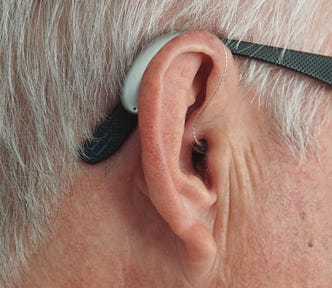 This image shows a close-up view of a person’s ear. The individual is wearing a behind-the-ear hearing aid in their right ear. The hearing aid is white, and there’s also a pair of glasses with a black frame visible, suggesting the individual may be older, as indicated by the gray hair.