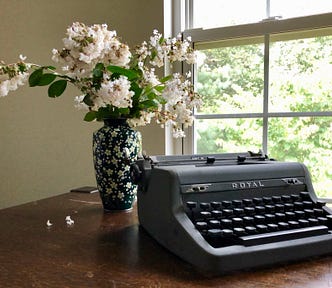 Image shows a brown tabletop in front of a window to the right of the image, and on the table an old typewriter and half behind it a vase with flowers.