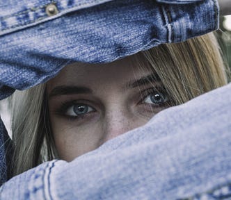 Close up of a young blonde in a jean jacket holding her arms up so just her eyes are visible