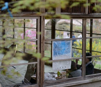 An old-style hard copy wall calendar is stuck to a window in an office facing out. We are outside, looking in at the calendar and the 2 people working at a desk in the background.