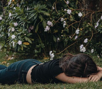 Prone young woman napping outdoors on a lawn by a bush.