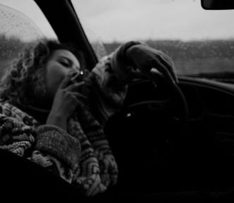 A black and white photo of a woman in a car wearing a sweater. She has a lit cigarette pressed to her lips. There is dew on the window and windshield. The mood is dark and dreary.
