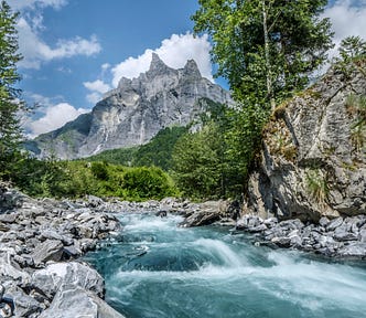 A fast-flowing river with trees and a mountain in the background, against a blue sky dotted with clouds.