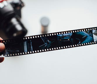 A film-negative strip from an analog camera. The camera is in soft focus in the background, with a canister of film next to it.