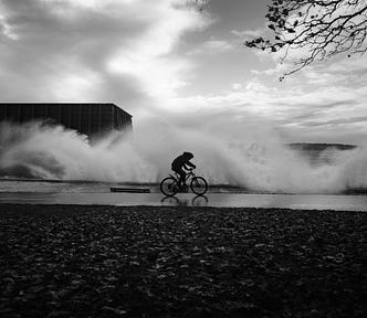 A cyclist rides along a wet road as waves crash over the sea wall behind, and the sun breaks through the clouds