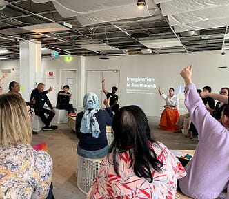 A group of people sit in a circle on stools with their thumbs up in the air, conveying that they think something is true.