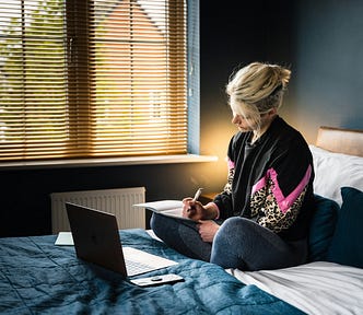 A woman sitting on her bed journaling, with a laptop in front of her in a dark blue bedroom.