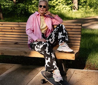 Elderly white woman with short white hair, big orange hoop earrings, and black sunglasses, wearing a pink button-down shirt with a yellow top underneath, with velvet black-and-white jersey cow patterned pants and white sneakers. She’s sitting on a wooden bench with one foot on a black skateboard, in partial sun at a park.