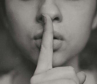 A womans face from the nose down, with an index finger up to the lip implying “be quiet”. Photo is black and white.