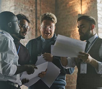Four people engaged in discussion while holding papers in a well-lit, industrial-style room with large windows.