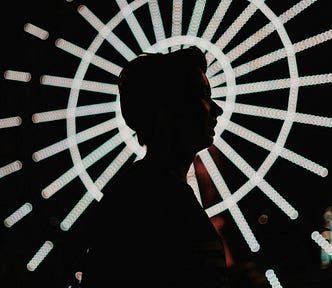 Person silhouetted against the lit spokes of a ferris wheel, as if the light is emanating from them