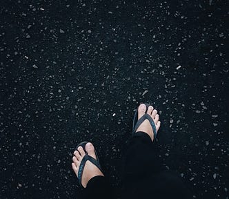 Black flip-flop wearing from knee down in black sweats, walking on black tar with plain feet.