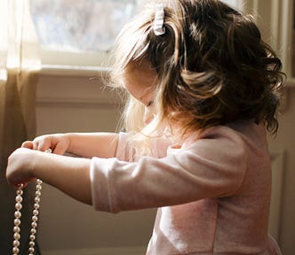 A side-view of a little girl, barely the height of the windowsill, wearing a pink dress holding out a string of pearls as if determining to put them around her neck. Her hair short and wavy but maturely coiffed like a woman’s but with a barrette. A representative photo of a girl growing up too fast against more idealistic fashion.