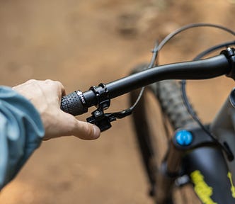 A photo of a man's left hand, reaching down to grasp the left handlebar of a bicycle. The handlebars are black, and we only see the left-hand side of the handlebars, with the front wheel of the bicycle out of focus in the background of the picture.