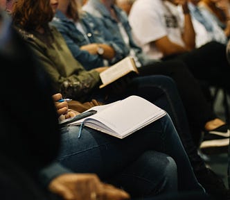 A group of people sit in a row of chairs, with notebooks open in their laps and pens in hand.