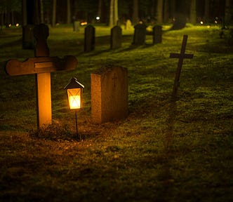 An image of several gravestones in a graveyard at night. A lantern glows gold in the foreground and lights the stones.
