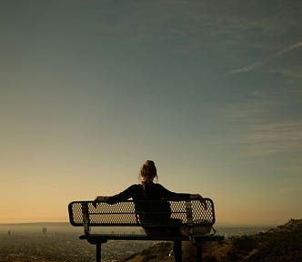 A young woman sits alone on a park bench, gazing forward as she faces away from us as the sun sets.