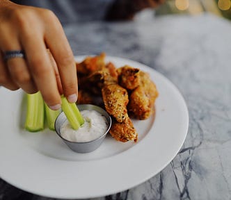 Close-up photo of a white plate with fried chicken tenders and celery, and a hand dipping some celery into a small metal bowl of ranch dressing.