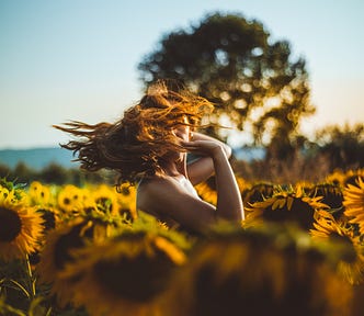 Woman with red hair standing in a field with sunflowers, her hair whipped in front of her face so that her face can’t be seen