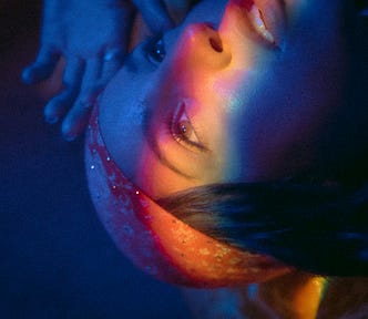 A blue toned image of a woman looking away. She wears a orange bandana and rainbow of light glows across her face.