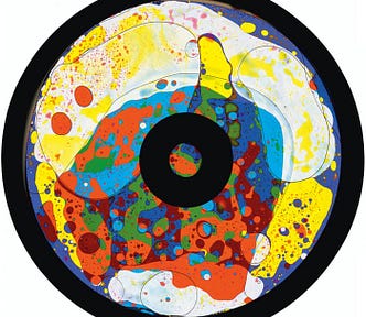 A photo of the “Cosmic Flare Oil Wheel” by Optikinetics. It is a round wheel, flat like a record in a record player, seen from above. It has multicolored splashes of paint/dye all over it in irregular patters — blobs of orange, areas of yellow and white, blue and red too. The periphery of the disk is dark black, as the center