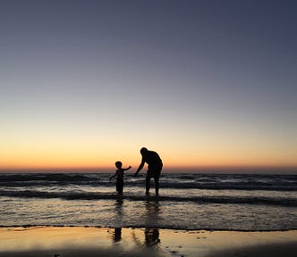 Two figures, of a child on the left and an adult on the right, are seen in silhouette on a beach as the sun sets.