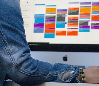 E-calendar view on Apple desktop. They are color-coded and time blocked. A person’s arm and hand are in the foreground — it appears that they’re scheduling an event on this calendar.