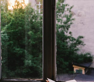 A book sits open on a window ledge, with a view of a building and trees beyond.