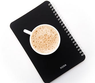 Black spiral-notebook for note taking with cappuccino cup sitting on the cover