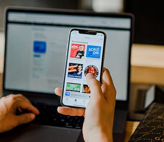 A person holding a smartphone with a list of ebook icons in one hand and typing on a laptop in the background with their other hand.