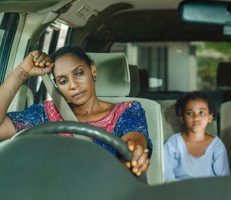 A mom, looking exhausted is sitting in her car, looking down, weighed down with trouble, her daughter sits in the back seat looking on