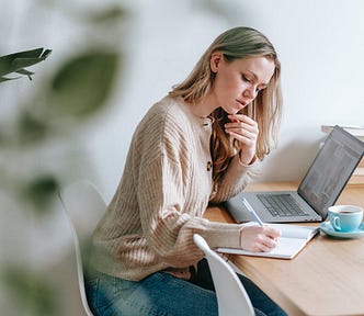 A woman is sitting at the table with her laptop open, writing something down in a notebook. #freelancewriter #workfromhome #writingopportunities #freelancewriting #writing #writer