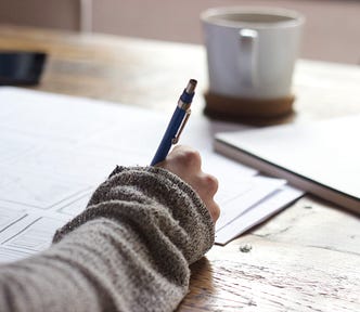 A writer sits at a desk, pen in sweatered hand, writing on large pieces of paper. A mug sits on a coaster above the work, with a notebook to the right of it.