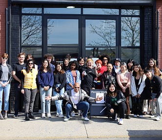 A group of 22 people are in front of a brick building with large glass doors. They stand around a storefront sign that reads “Pioneer Works. Code, Decolonized with Power Plant” and smile into the camera.