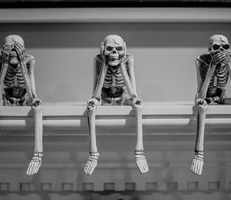 Three skeletons sitting on a ledge in the “see no evil, hear no evil, speak no evil” positions.