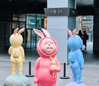 Three statues of what could be Rabbit Man outside of an office building. One dressed in yellow, one in a pinkish/red, and the third in blue. All three are looking skyward wondering “What the bloody hell is going on?”