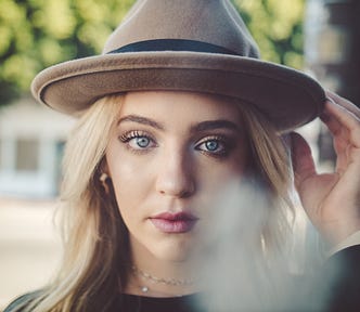 Close up of a young blonde woman with bright blue eyes, holding her hand up to the brim of her beige hat