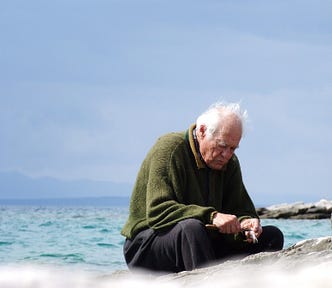 Old man sitting on a beach and looking down, appearing to be sad.