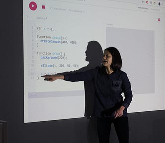 Asian-American woman with long dark hair stands in front of a projection of p5.js editor.