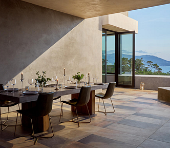 Modern, minimalist terrace at sunset: a long concrete dining table set for six with candles, glassware, and greenery, surrounded by sleek black chairs on a tiled floor. An open wall frames a distant ocean-and-mountain view, and a square gas fire pit glows on the right.